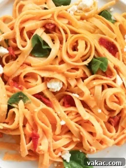 A photo of creamy red pepper pasta on a white plate. Ingredients include: fettuccine noodles, red pepper, hummust, spinach, sundried tomatoes, feta cheese.