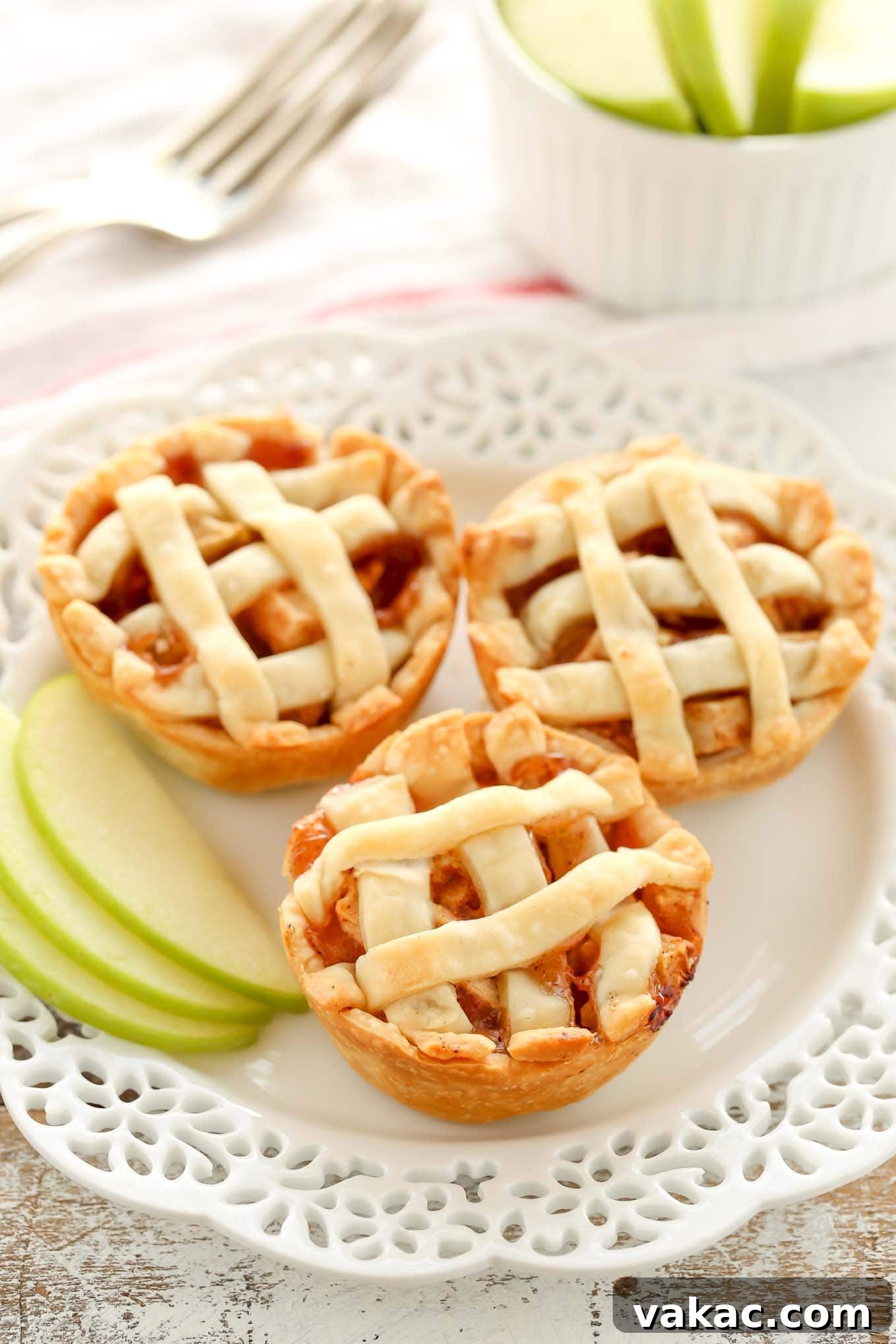 Sweet Apple Pie Bites 2 Three mini apple pies on a decorative white plate. A small container of sliced apples and a fork are in the background.