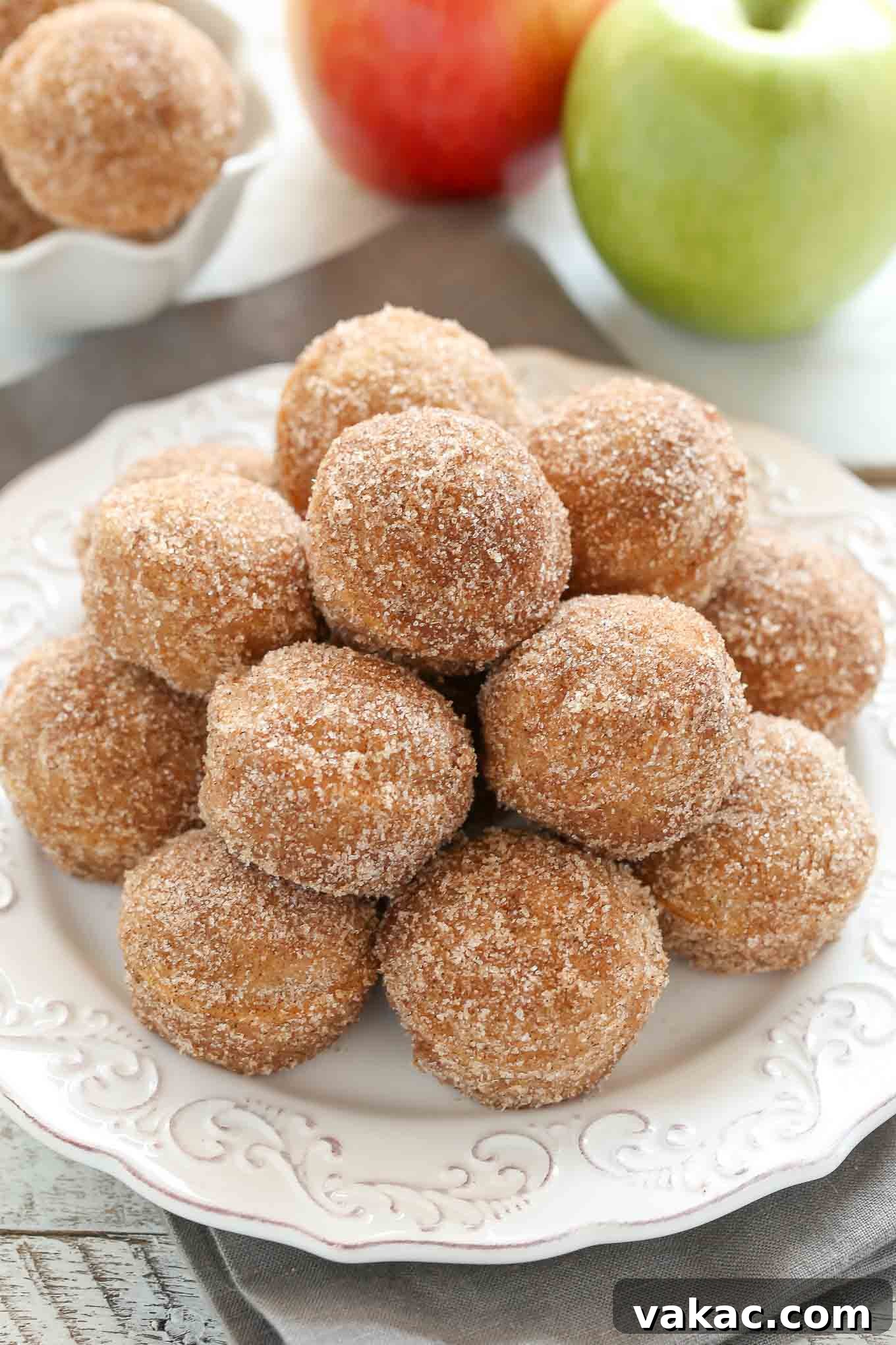 Baked Apple Cider Donut Bites 2 A white plate piled high with golden-brown baked apple cider donut holes. Two fresh apples, one red and one green, rest blurred in the background, hinting at autumn.