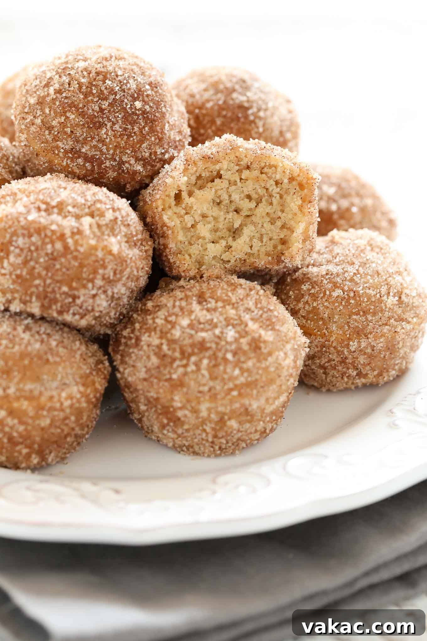 Baked Apple Cider Donut Bites 3 Close-up view of a pile of freshly baked apple cider donut holes on a white plate. The donut hole in the center has a bite taken out of it, revealing its soft, moist interior.