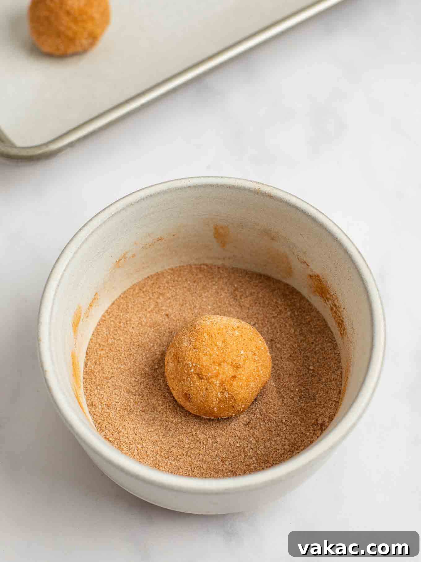 Spiced Pumpkin Snickerdoodles 8 A ball of pumpkin snickerdoodle cookie dough is being rolled meticulously in a mixture of cinnamon and sugar, preparing it for baking.