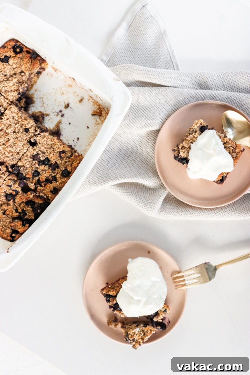 Blueberry baked oatmeal in a white dish cut up and served onto two pink plates topped with Greek Yogurt. Ingredients include: oats. blueberries, cinnamon