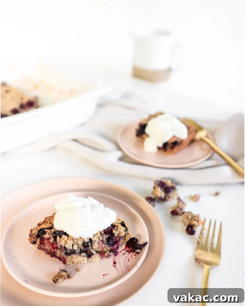 Blueberry baked oatmeal served onto two pink plates topped with Greek Yogurt. Ingredients include: oats, banana, blueberries, cinnamon, maple syrup.