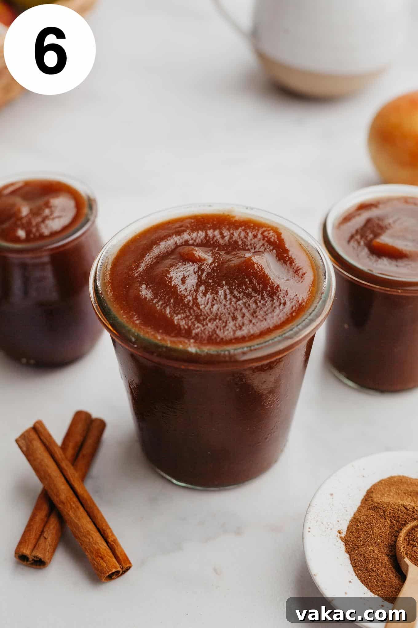 Classic Homemade Apple Butter Recipe 9 Three glass jars of homemade apple butter arranged on a wooden counter, garnished with cinnamon sticks and scattered whole spices, creating an inviting autumnal scene.