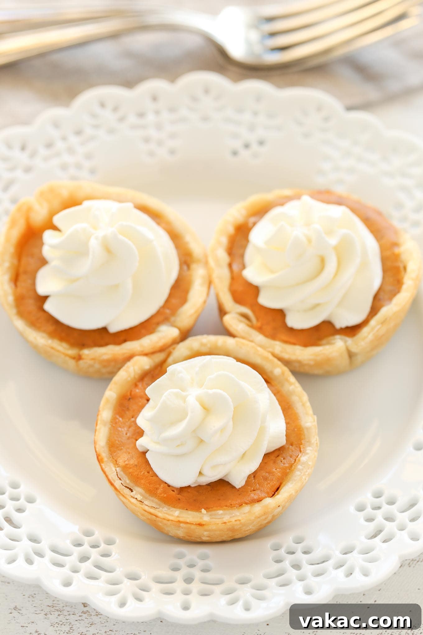 Pumpkin Pie Bites 2 An angled overhead view of three mini pumpkin pies topped with whipped cream on a white plate. Two forks rest in the background.