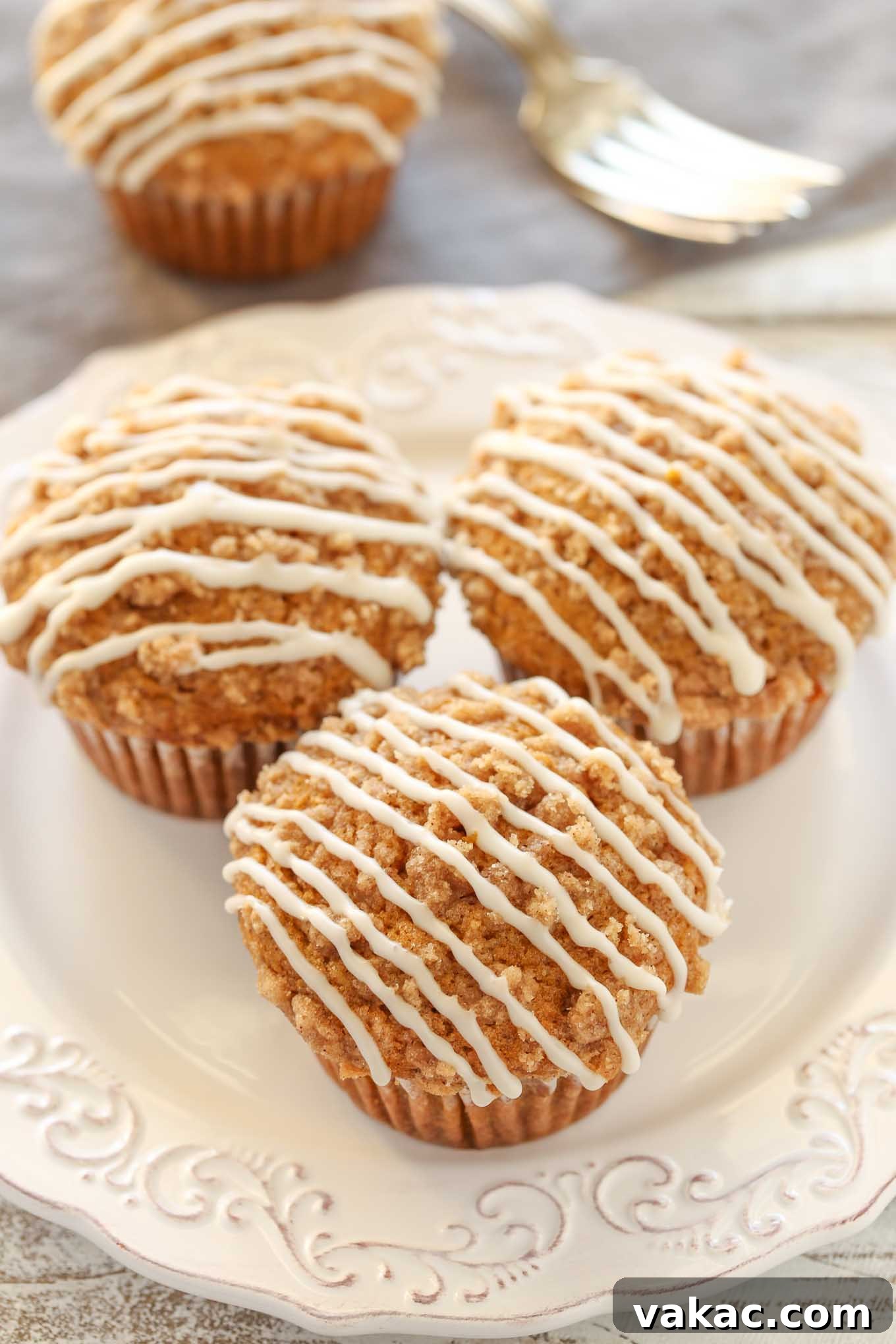 Three glazed pumpkin streusel muffins on a white plate. Another muffin and a fork rest in the background. 