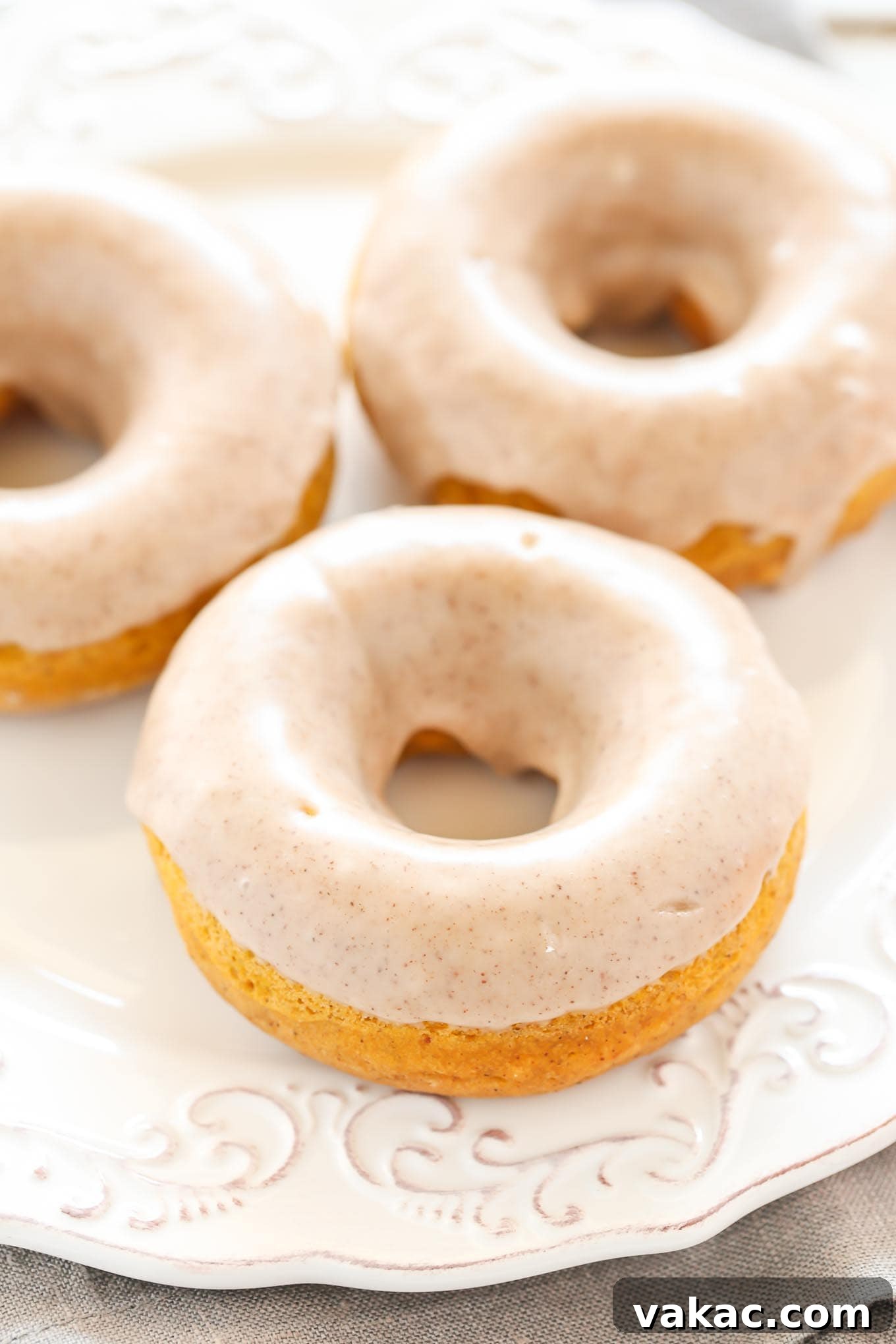 Cozy Autumn Pumpkin Doughnuts with Maple Cinnamon Drizzle 3 Three golden-brown baked pumpkin donuts, generously coated with a rich maple cinnamon glaze, rest invitingly on a pristine white plate. A subtle drizzle of glaze decorates the plate, hinting at their deliciousness.