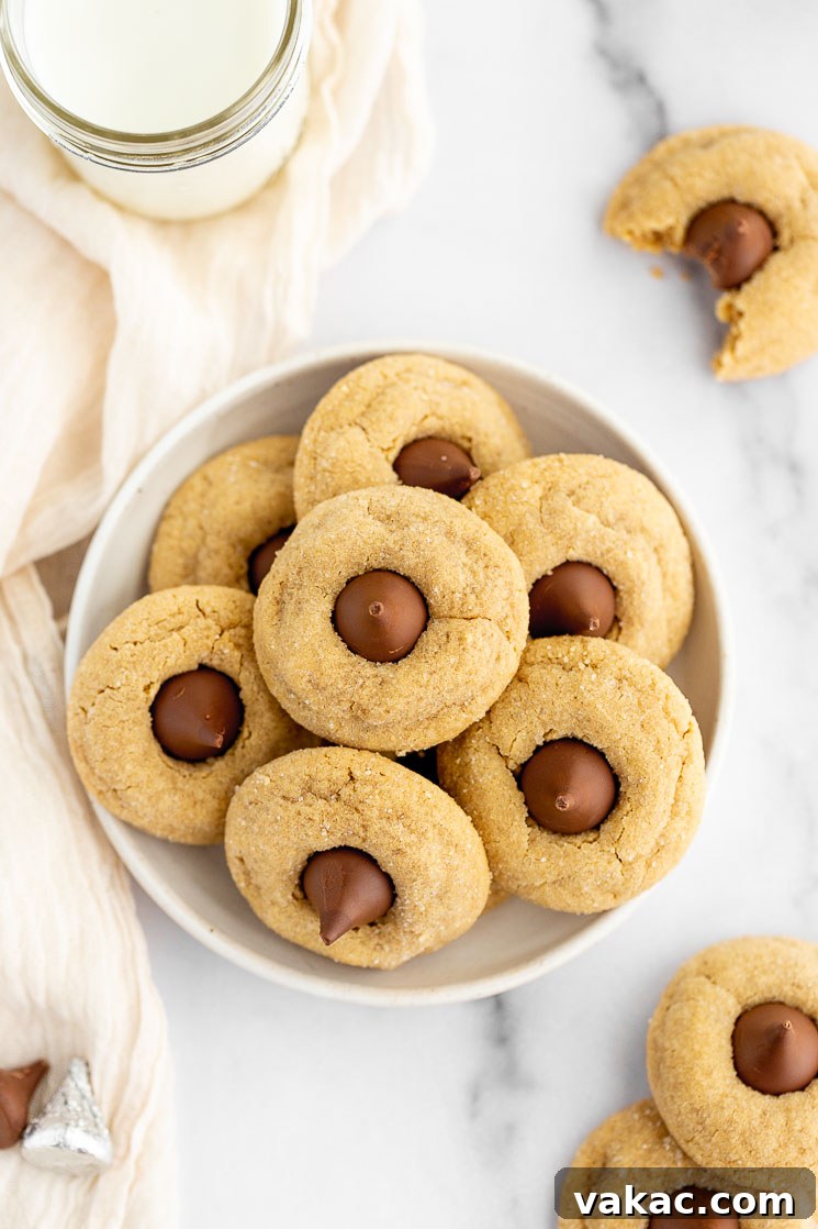 A plate with several peanut butter blossom cookies stacked on it with a glass of milk and a cookie with a bite taken out of it in the background.