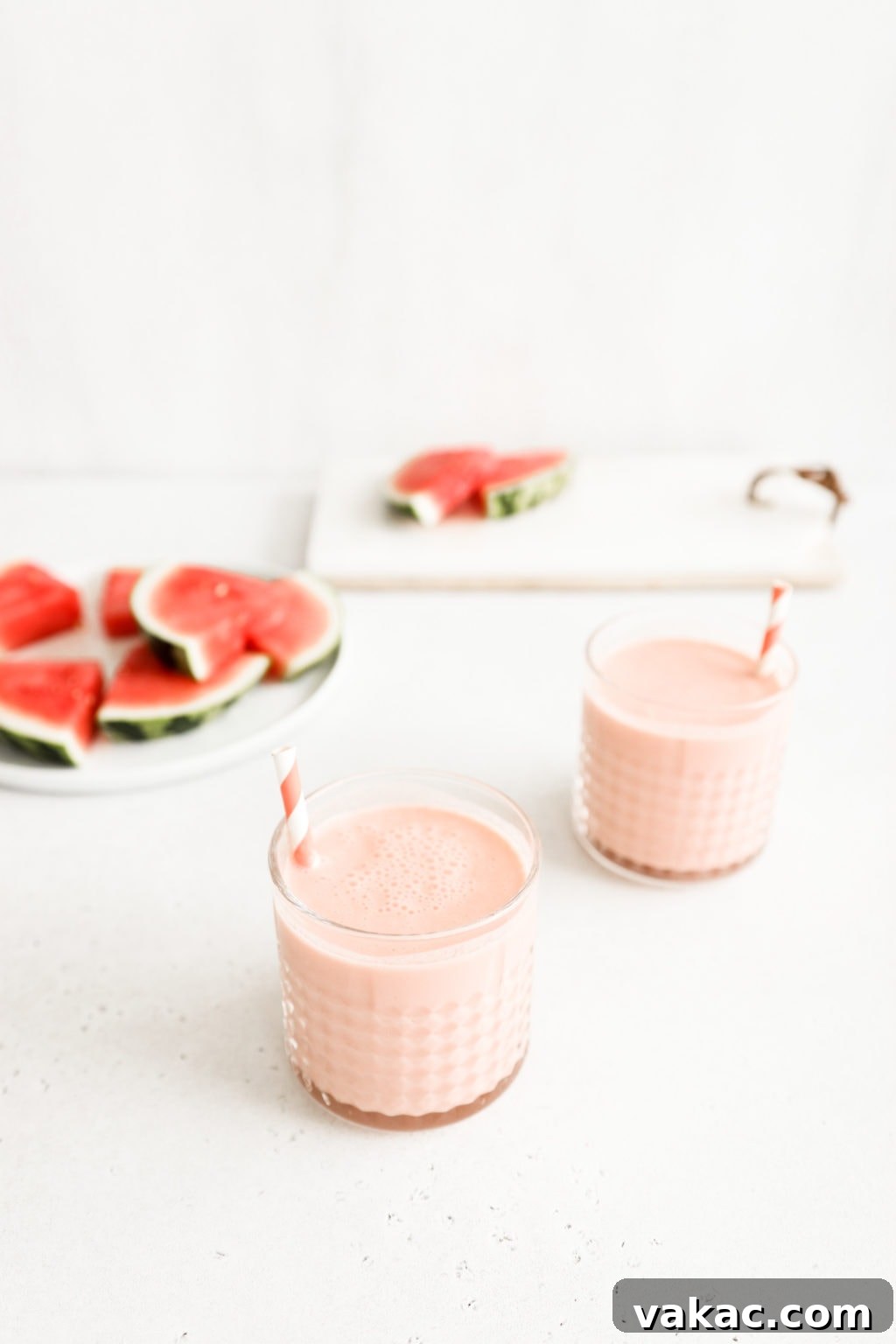 Refreshing 5-Minute Watermelon Banana Bliss 4 Two vibrant watermelon banana smoothies in elegant glass cups, each adorned with a pink and white striped straw. They are arranged on a clean, neutral surface, with artistic blurred slices of fresh watermelon in the background, highlighting the key ingredients.