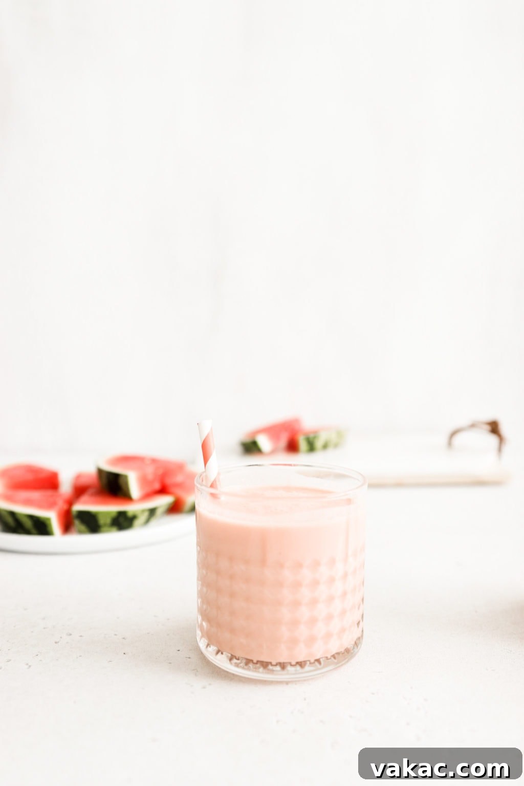 Refreshing 5-Minute Watermelon Banana Bliss 5 A single, beautifully blended watermelon banana smoothie in a clear glass cup, featuring a festive pink and white striped straw. The glass rests on a clean, neutral surface, with artistic, out-of-focus slices of fresh watermelon creating a vibrant backdrop.
