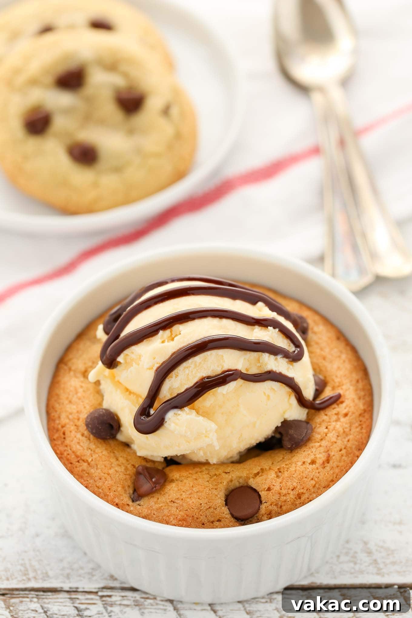 A deep dish single serve chocolate chip cookie topped with ice cream and chocolate sauce. Two cookies rest on a plate in the background.