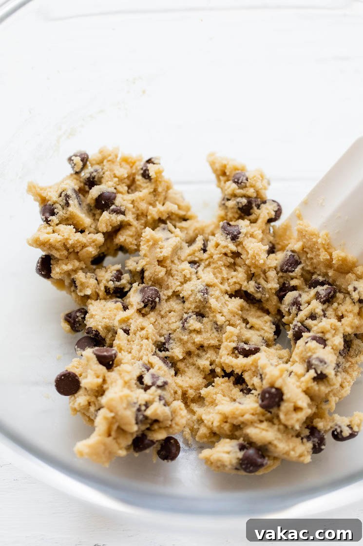 Mini Chocolate Chip Wonders 4 Cookie dough being gently mixed with a white spatula in a clear glass bowl, showcasing the rich texture before baking.