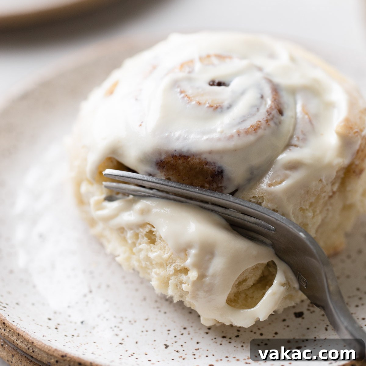 A single cinnamon roll on a white speckled plate being cut into with a fork.