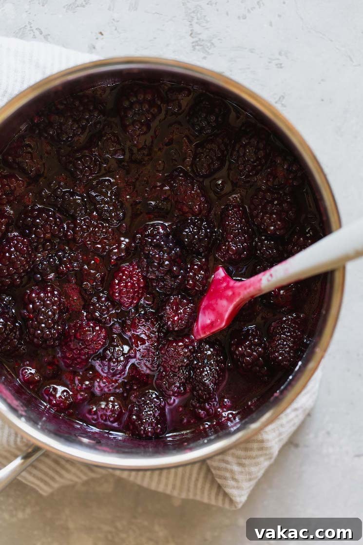 A saucepan filled with sweet blackberry filling being stirred gently with a white spatula, showing the berries softening and juices thickening.