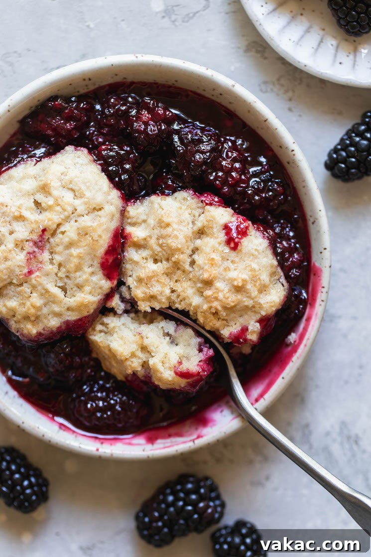 An antique bowl generously filled with warm blackberry cobbler, featuring a spoon taking a bite out of the biscuit topping.