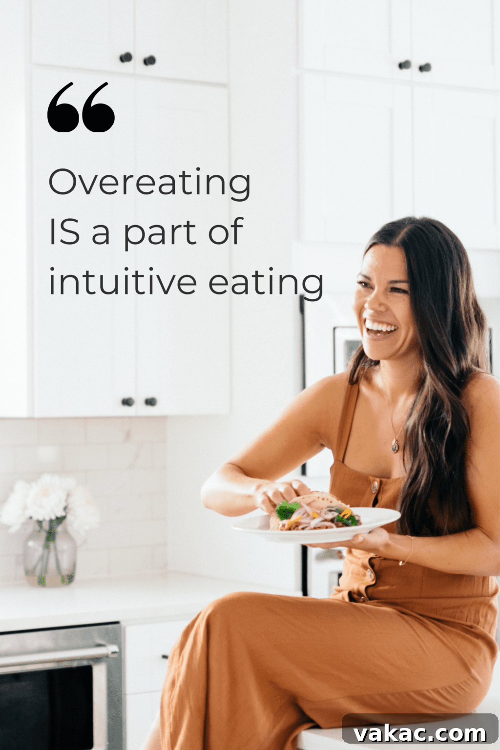 A woman with black hair is sitting on a counter in a brown dress. She is in a white kitchen eating a taco. There is text overlay to the left of the girl that reads "overeating is a part of intuitive eating".