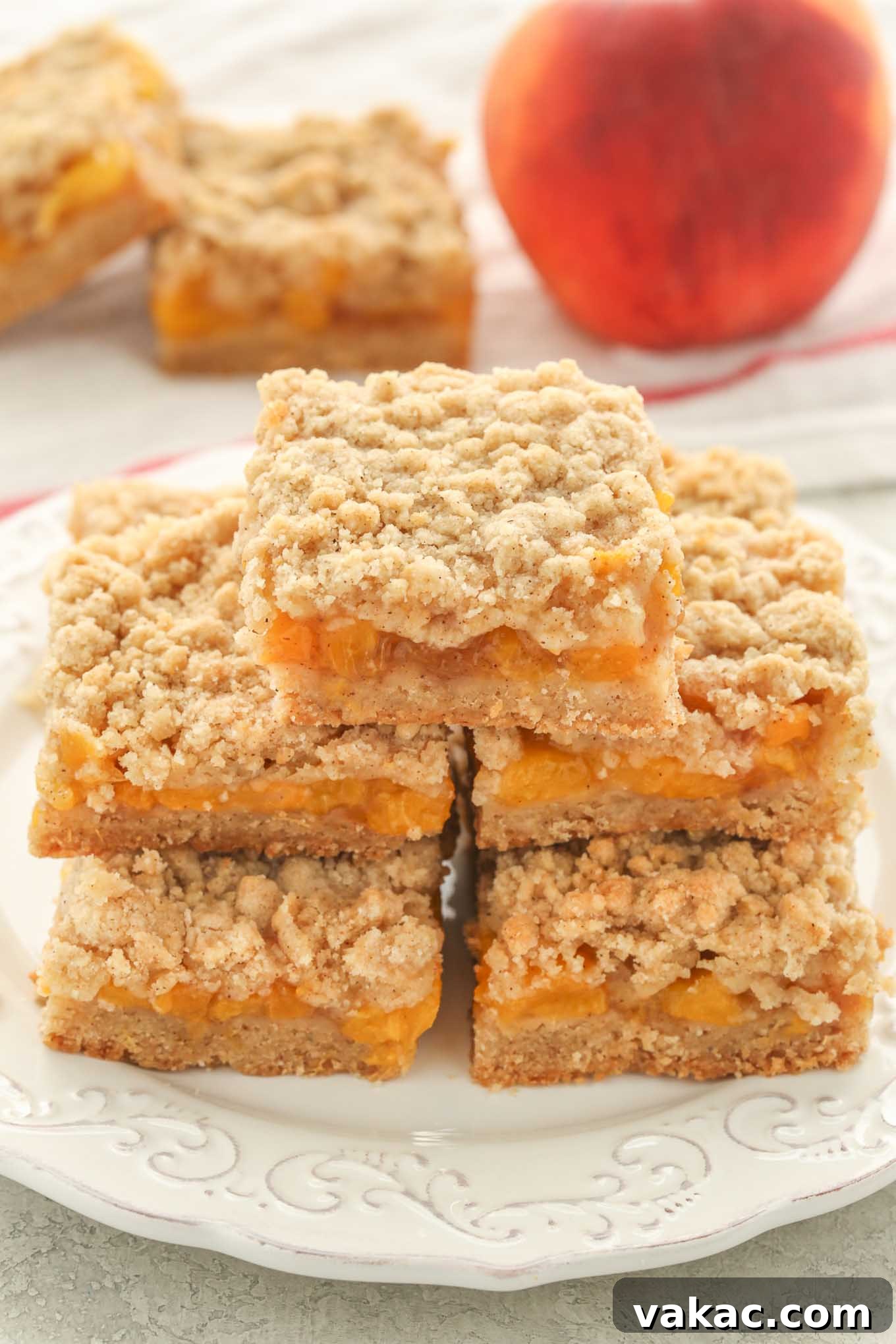 Sun-Kissed Peach Crumble Bars 5 A delightful pile of perfectly baked peach crumble bars arranged on a pristine white plate, with a fresh peach and two more bars in the soft-focus background.