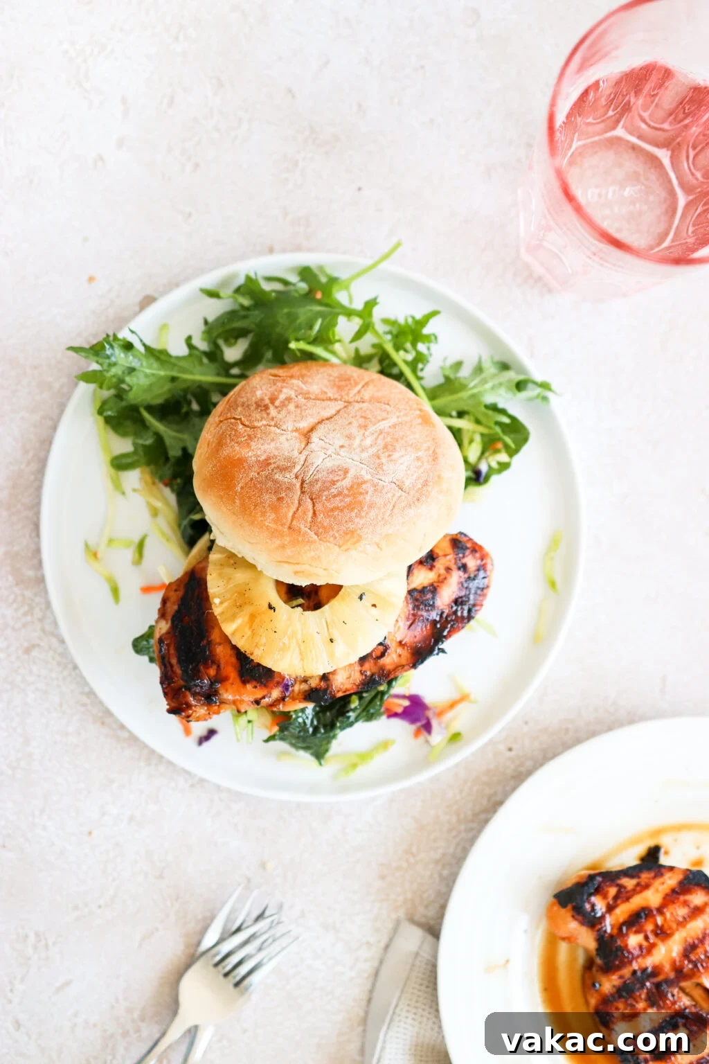 Sweet & Umami Maple-Soy Chicken Burger 2 A grilled chicken burger topped with a pineapple ring is on a white plate and is surrounded by an arugula salad.