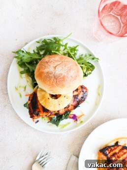 A grilled chicken burger topped with a pineapple ring is on a white plate and is surrounded by an arugula salad.