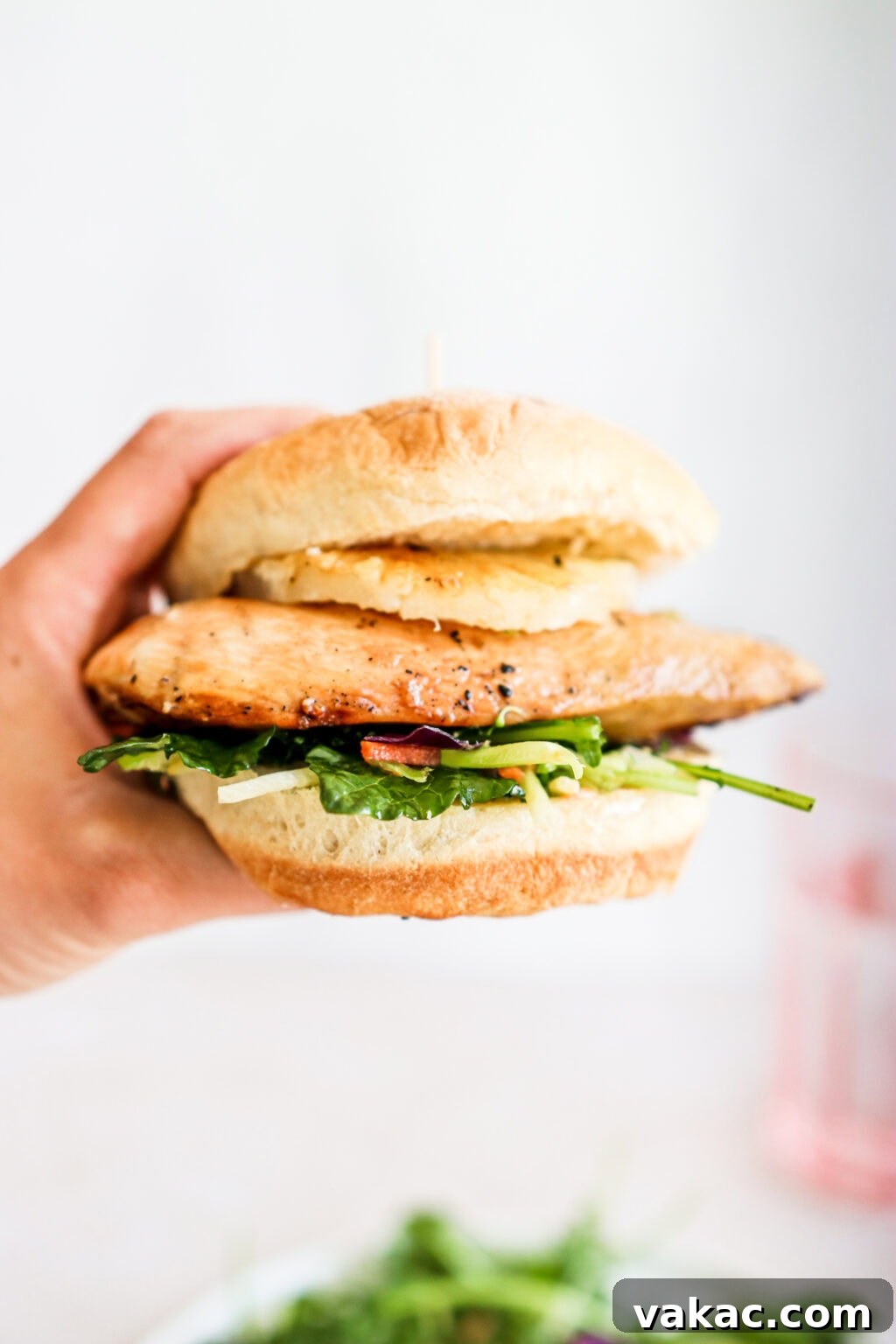 Sweet & Umami Maple-Soy Chicken Burger 9 a hand holding a burger bun layered with greens, slaw mix, maple soy glazed chicken breast and a pineapple ring with a pink glass in the background
