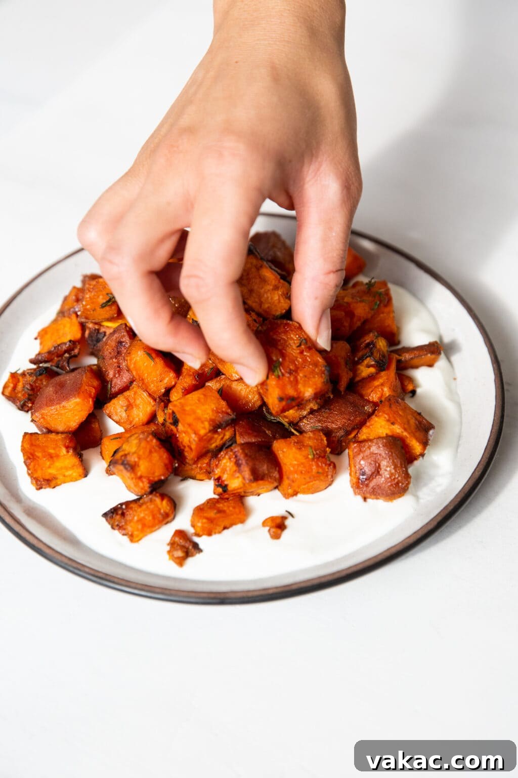 Irresistible Golden Sweet Potatoes with Velvety Garlic Aioli 5 A hand is picking up a baked sweet potato cube on a white plate with a brown rim.
