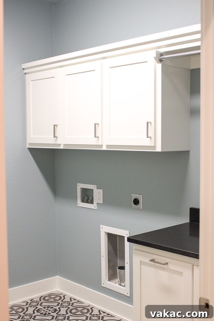 Our New Home Tour and Life Lately 16 A modern laundry room with serene blue-gray walls, sleek white cabinets, and durable gray porcelain tiles mimicking concrete, awaiting a washer and dryer.