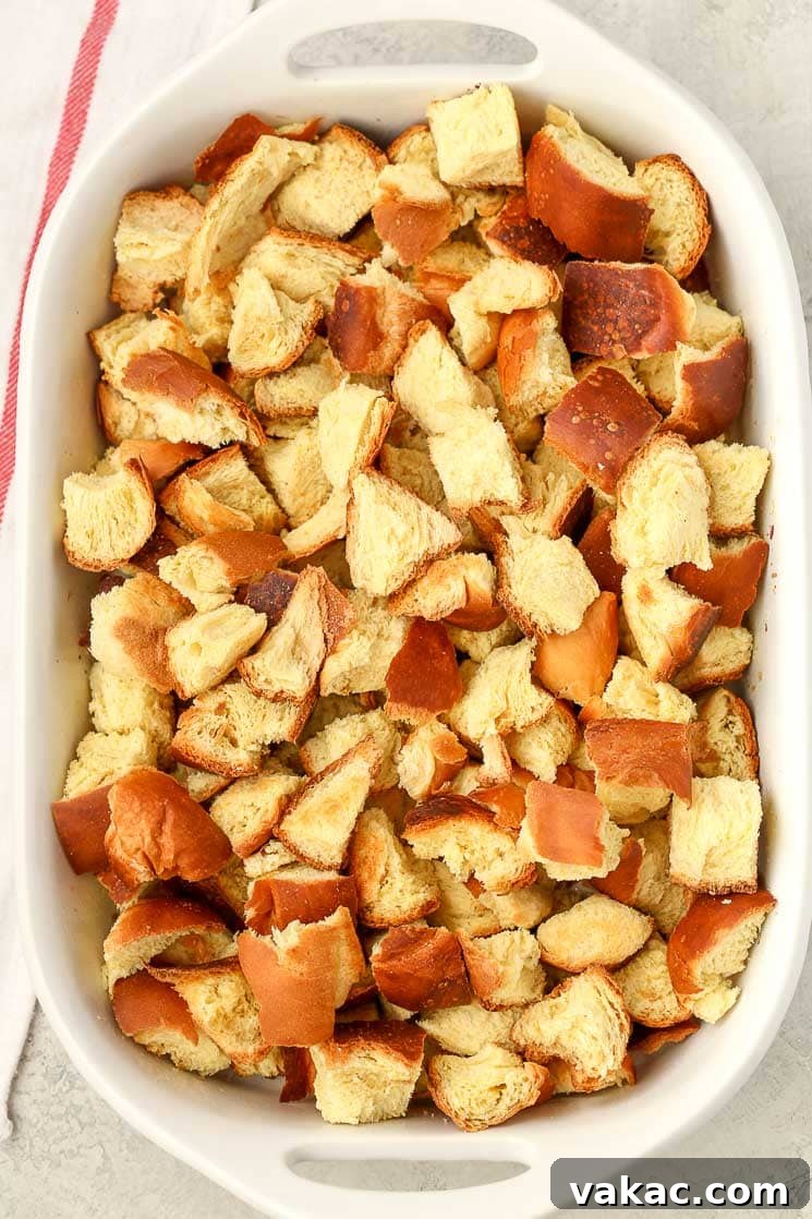 An overhead view of golden, cubed challah bread evenly spread in a white ceramic baking dish, ready for the next step in the bread pudding recipe.