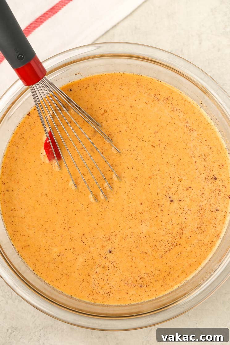 A glass bowl filled with the creamy, orange-hued wet ingredients for homemade bread pudding, with a whisk resting inside, ready for mixing.