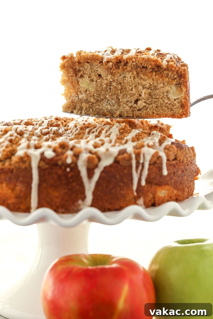 Apple Cinnamon Coffee Cake 4 A slice of apple streusel coffee cake being lifted from a cake stand. Two apples rest in the foreground.