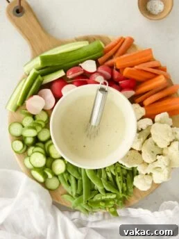 A wooden board with sliced carrots, radishes, cucumbers, cauliflower, and snap peas arranged around a bowl of creamy Caesar dressing with Greek yogurt and a small whisk, next to a small dish of salt.