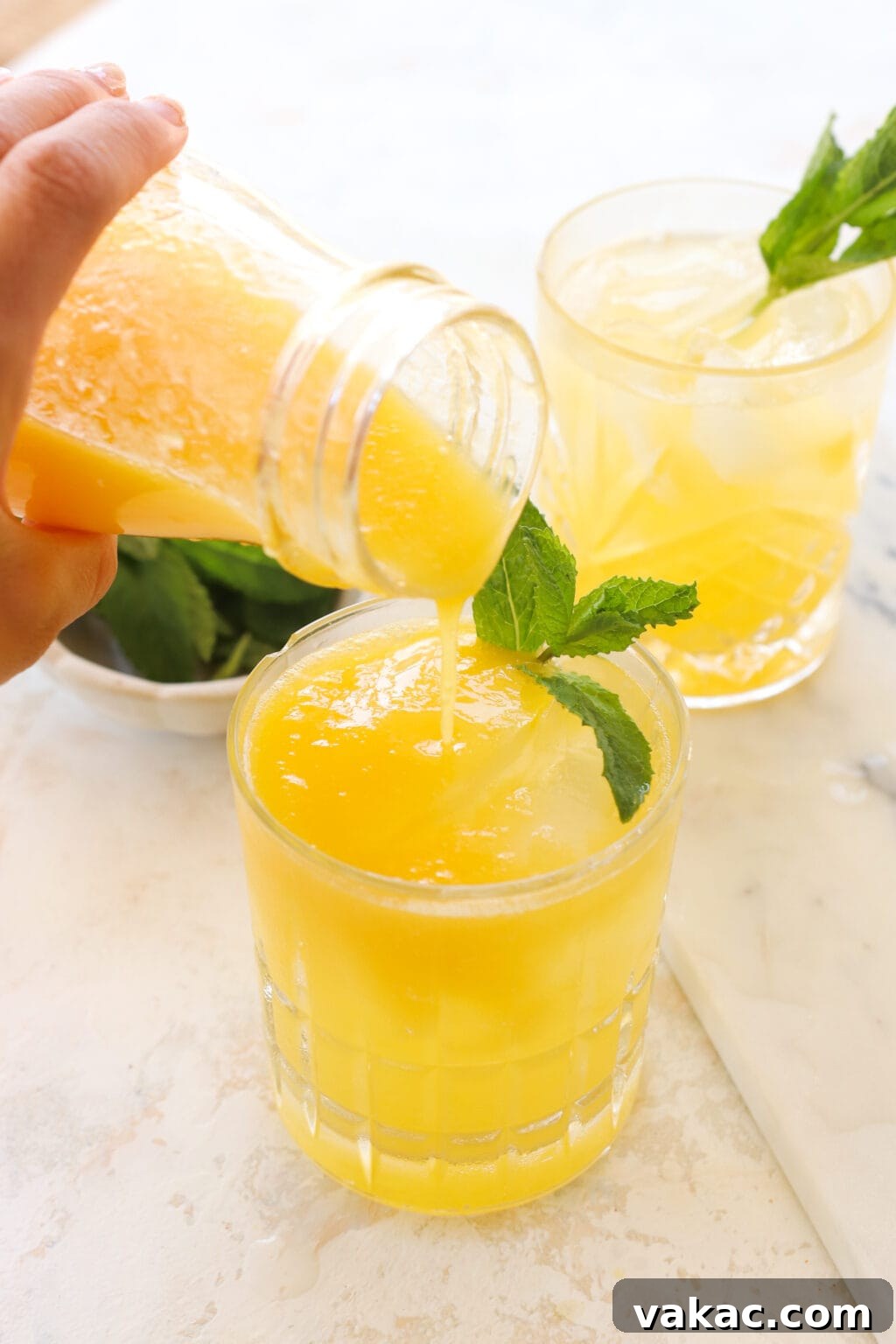 Effortless Wholesome Mango Puree 6 A hand pouring yellowish-orange mango puree from a bottle into a short clear glass garnished with mint. Another glass and a bowl of mint are blurred in the background.