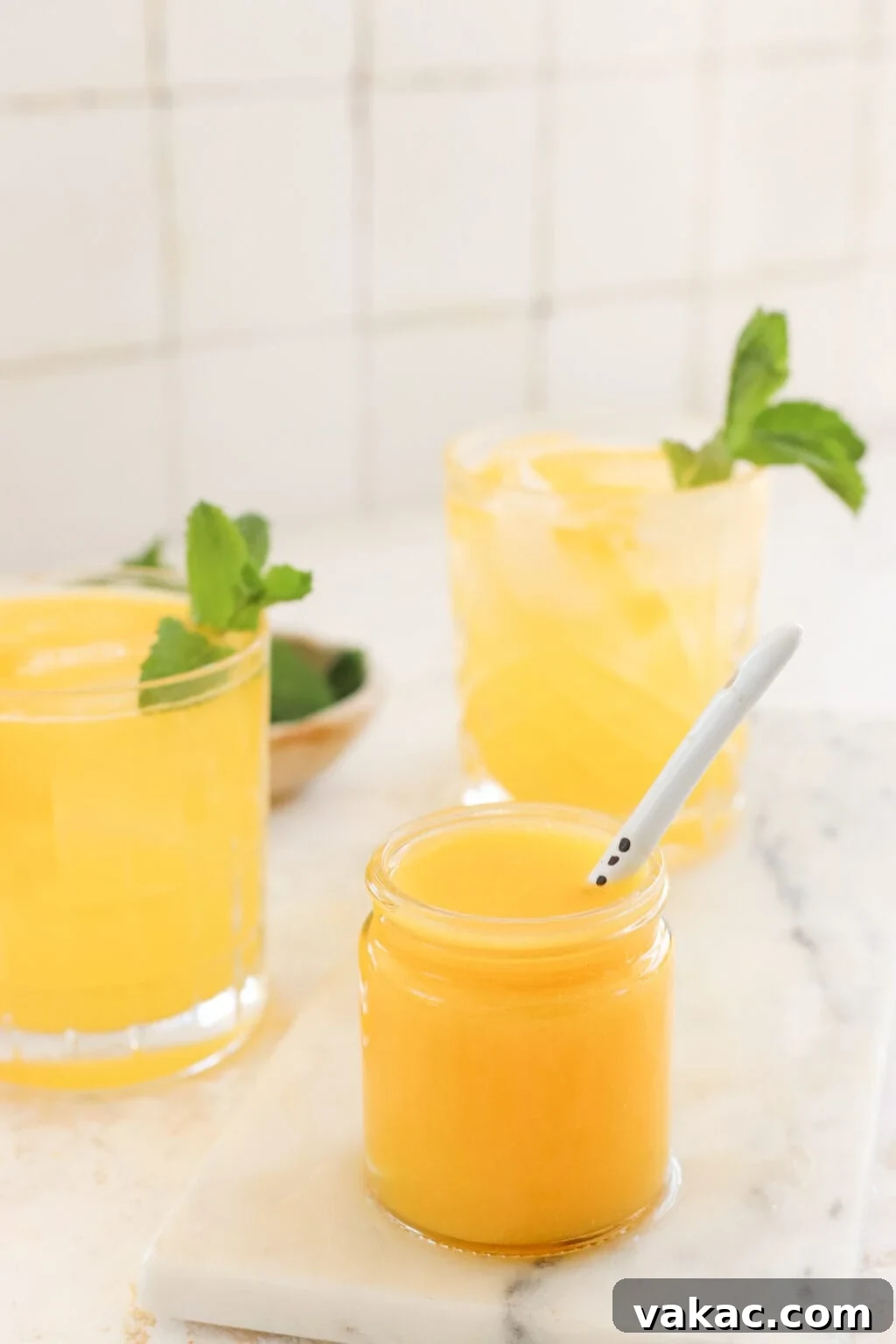 Effortless Wholesome Mango Puree 7 Two glasses with yellowish-orange liquid and mint garnishes stand in front of a white tile wall. A smaller jar with mango puree and a white spoon is placed in front of them.