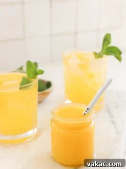 Two glasses with yellowish-orange liquid and mint garnishes stand in front of a white tile wall. A smaller jar with mango puree and a white spoon is placed in front of them.