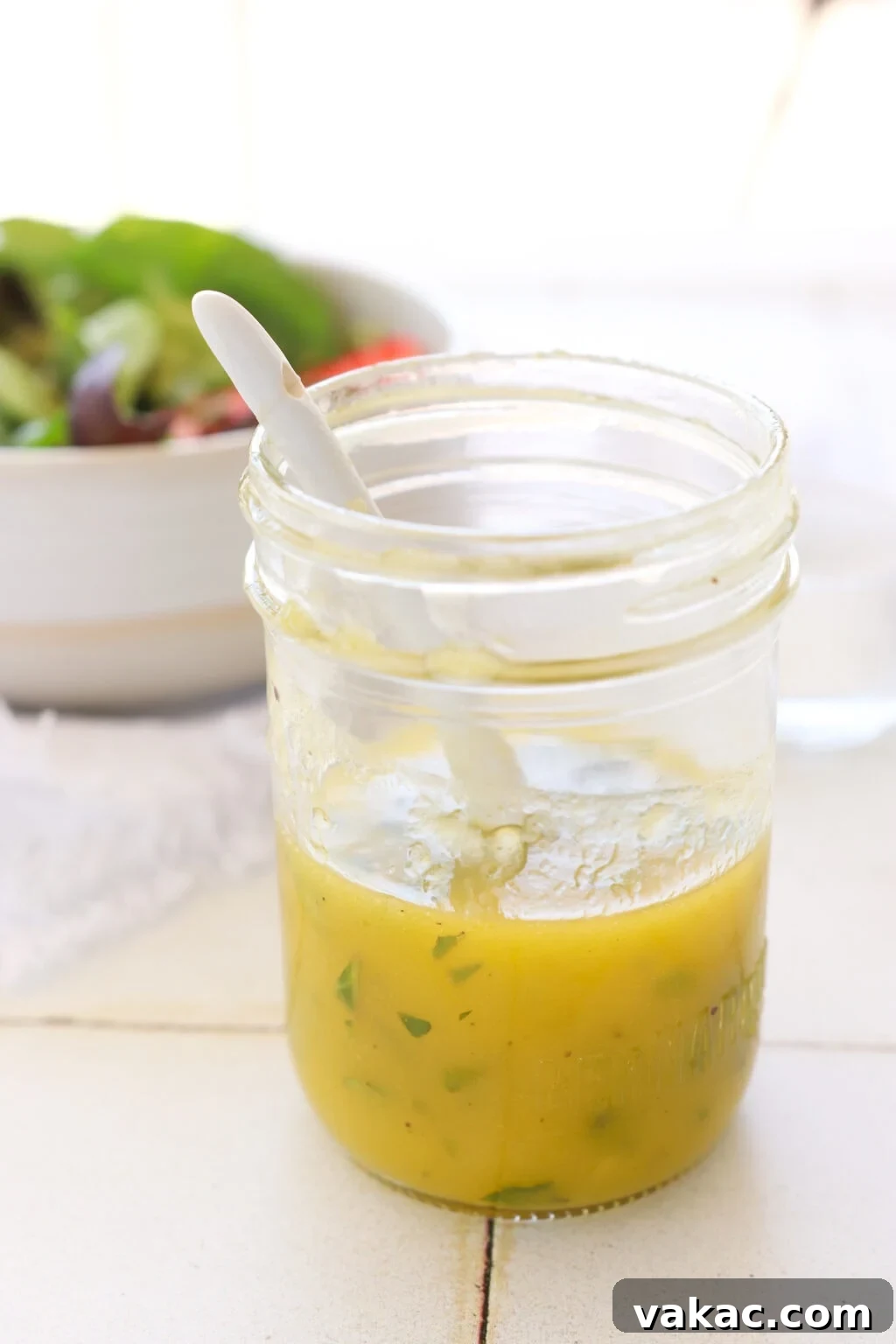 Zesty 5-Minute Mango Mint Vinaigrette 2 A mason jar with a bright yellow mango mint vinaigrette, a white spoon rests on a tiled counter. In the background, a colorful salad in a white bowl awaits.