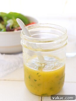 A mason jar with a bright yellow mango mint vinaigrette, a white spoon rests on a tiled counter. In the background, a colorful salad in a white bowl awaits.