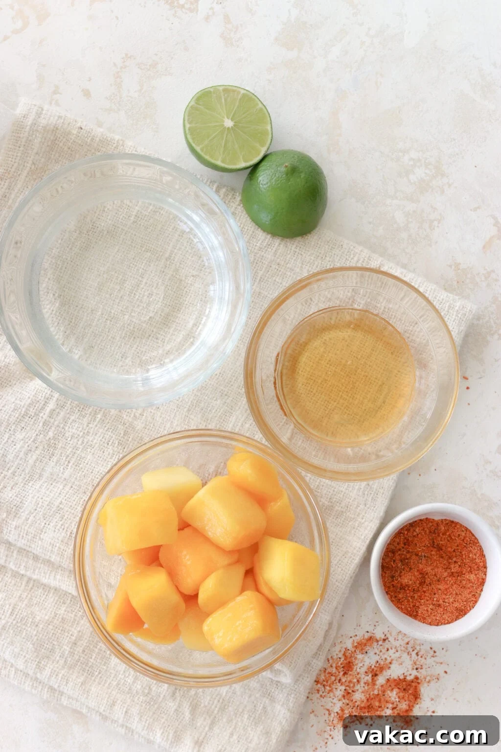 Sweet Heat Mango Tajin Popsicles 3 A beautifully arranged collection of ingredients for mango tajin popsicles sits on a clean white counter. Halved limes, a bowl of fresh honey, a bowl of frozen mango chunks, and a small dish of red Tajin seasoning are artfully displayed, ready for preparation.