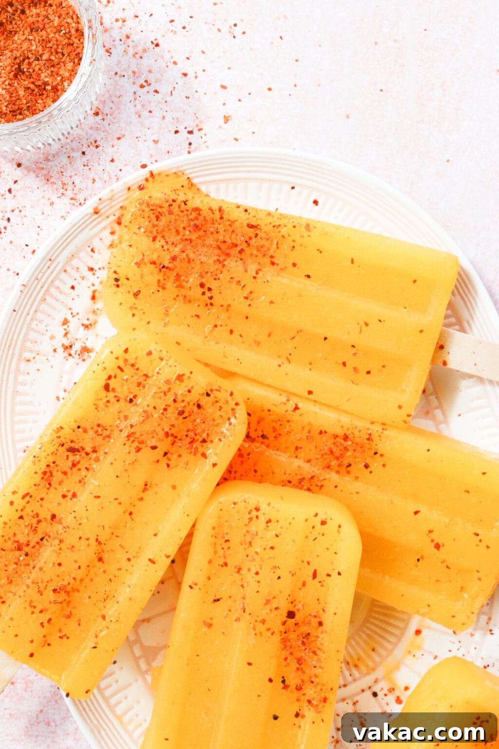 Sweet Heat Mango Tajin Popsicles 6 Four perfectly orange-yellow colored popsicles are arranged on a stylish pink and white oval plate. Each popsicle is beautifully sprinkled with the vibrant red Tajin seasoning, making them look even more appetizing.
