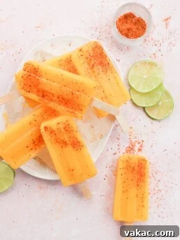 Four vibrant orange-yellow popsicles, generously coated with Tajin seasoning, rest on a charming white and pink plate, surrounded by fresh lime slices. Another popsicle lies invitingly in the corner.