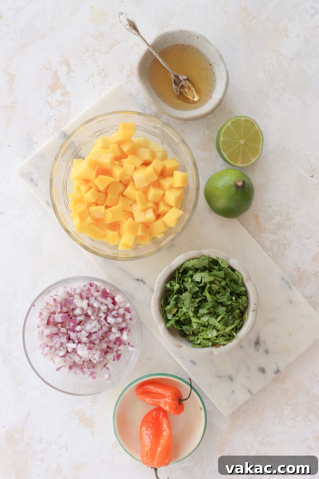 An overhead shot of ingredients to make mango habanero salsa. At the top in a grey dish is honey, below is a clear bowl with diced mango and two halves of lime. Below to the left is a small clear bowl with minced red onion and to the right is roughly chopped cilantro. At the bottom is a clear bowl with whole orange habanero peppers.  