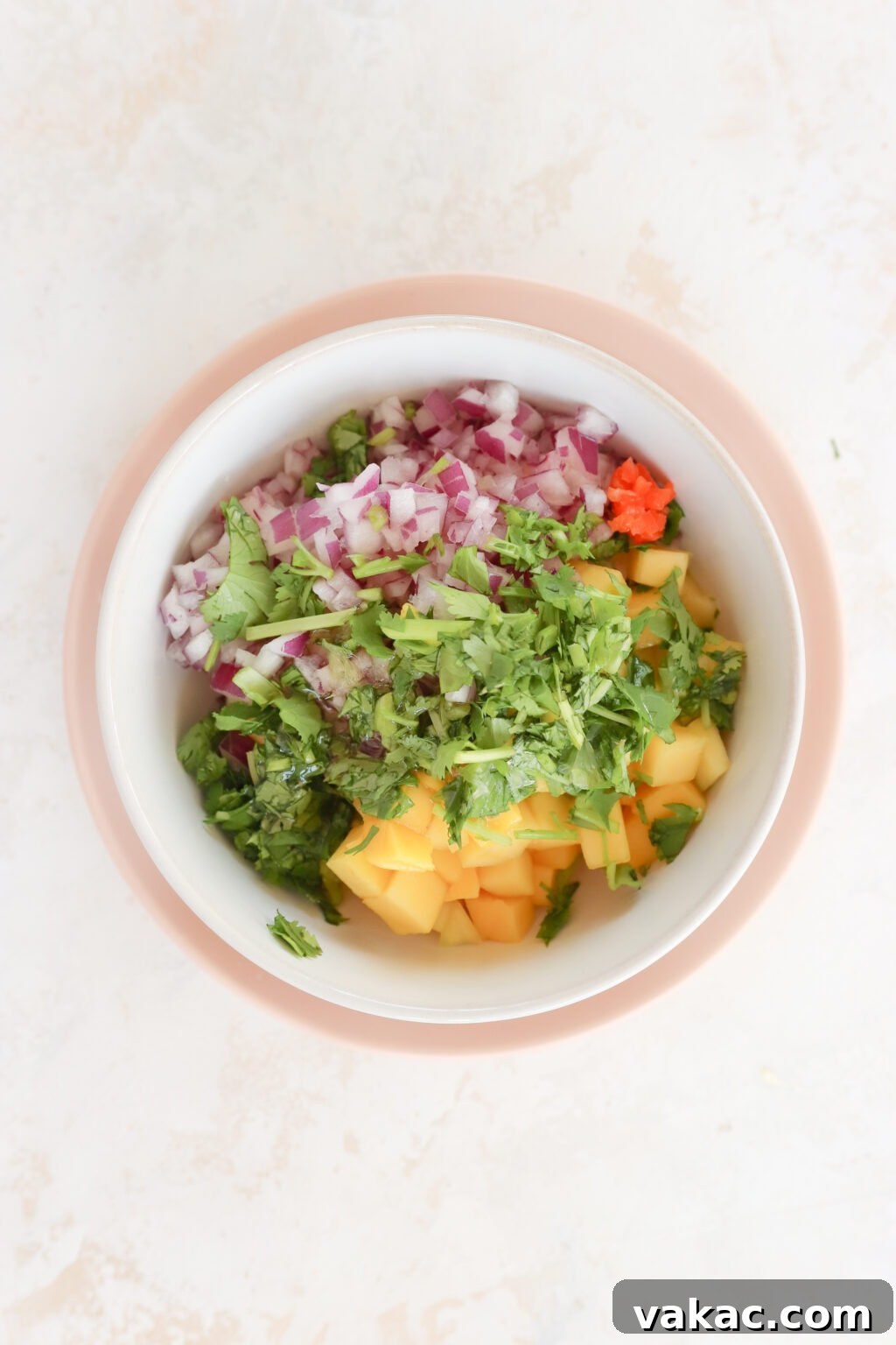 In a white bowl with a pink rim is ingredients for a salsa. There is cubed mango, diced red onion, diced habanero pepper, honey and cilantro all piled. The ingredients have not yet been mixed together.