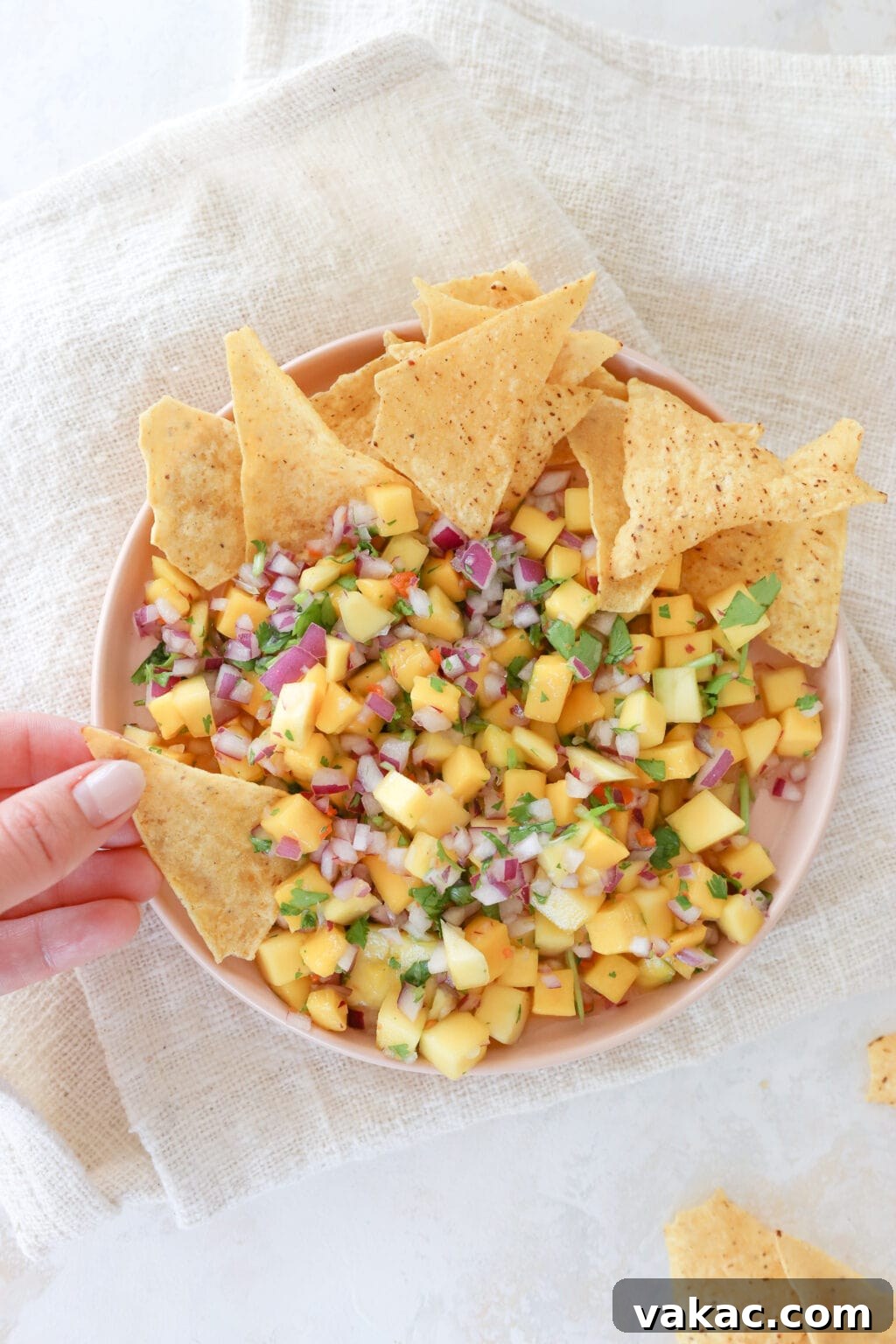 An overhead shot of a pink plate with a rim on top of a beige burlap style napkin. On the plate is a mixed mango salsa that has cubed mango, minced red onion, minced habanero pepper, and cilantro leaves. At the top of the plate is a stack on tortilla chips. A woman's hand is holding a chip and dipping it in the salsa.