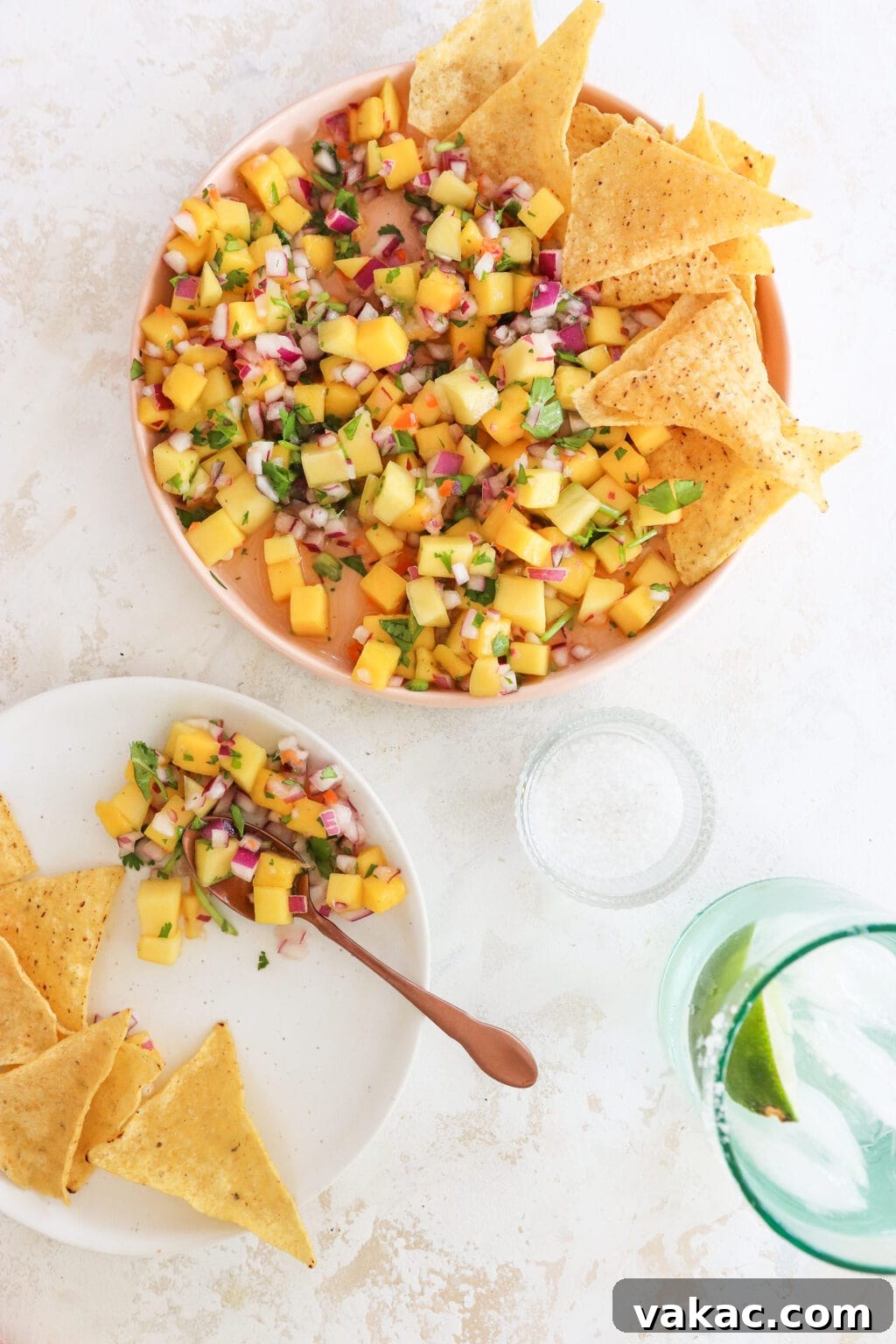 An overhead shot of a white counter top with a pink plate with a rim topped with salsa and chips. In the bottom left corner is a white plate with a copper spoon holding mango salsa and tortilla chips. In the bottom right corner is a greenish cup with soda water and lime.