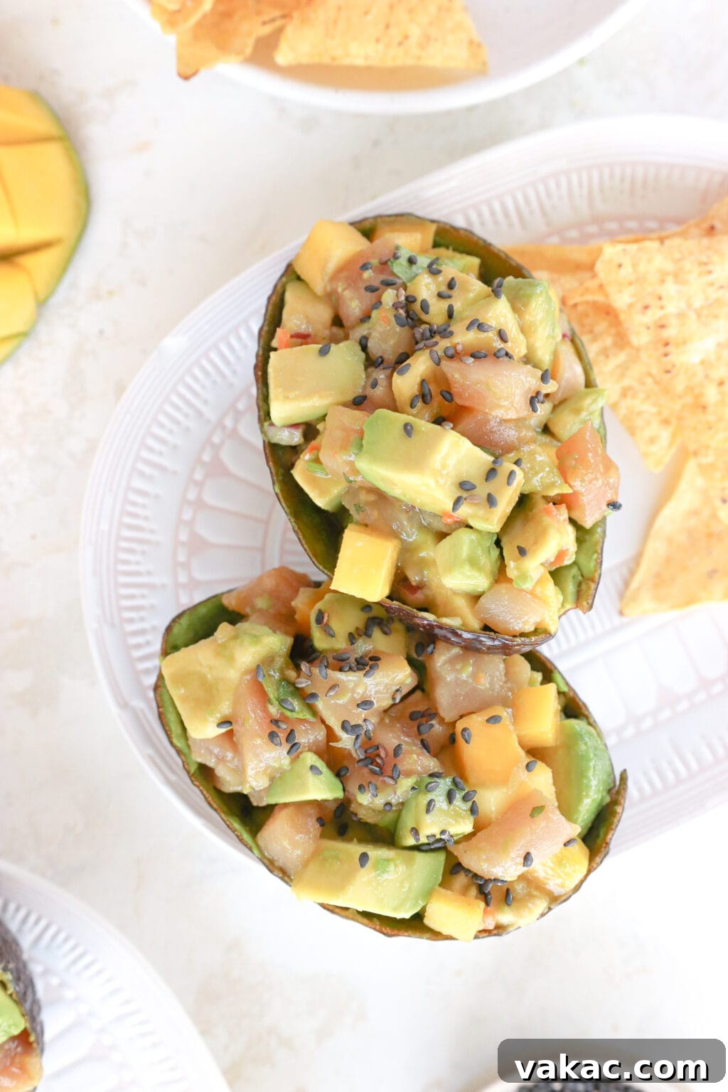 2 emptied avocado shells are stuffed with cubed raw tuna, mango, and avocado and are topped with black sesame seeds. They're on a white and pink plate with chips