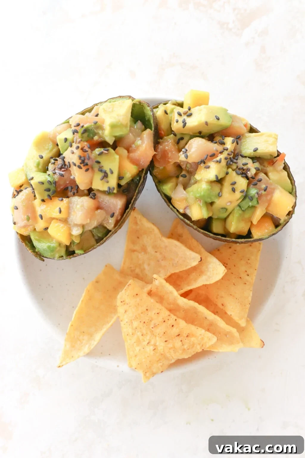 Two avocados are on a white plate. The avocados are stuffed with cubed avocado, cubed raw tuna, mango habanero salsa and topped with black sesame seeds. Below them on the plate are tortilla chips.