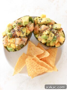 Two avocados are on a white plate. The avocados are stuffed with cubed avocado, cubed raw tuna, mango habanero salsa and topped with black sesame seeds. Below them on the plate are tortilla chips.