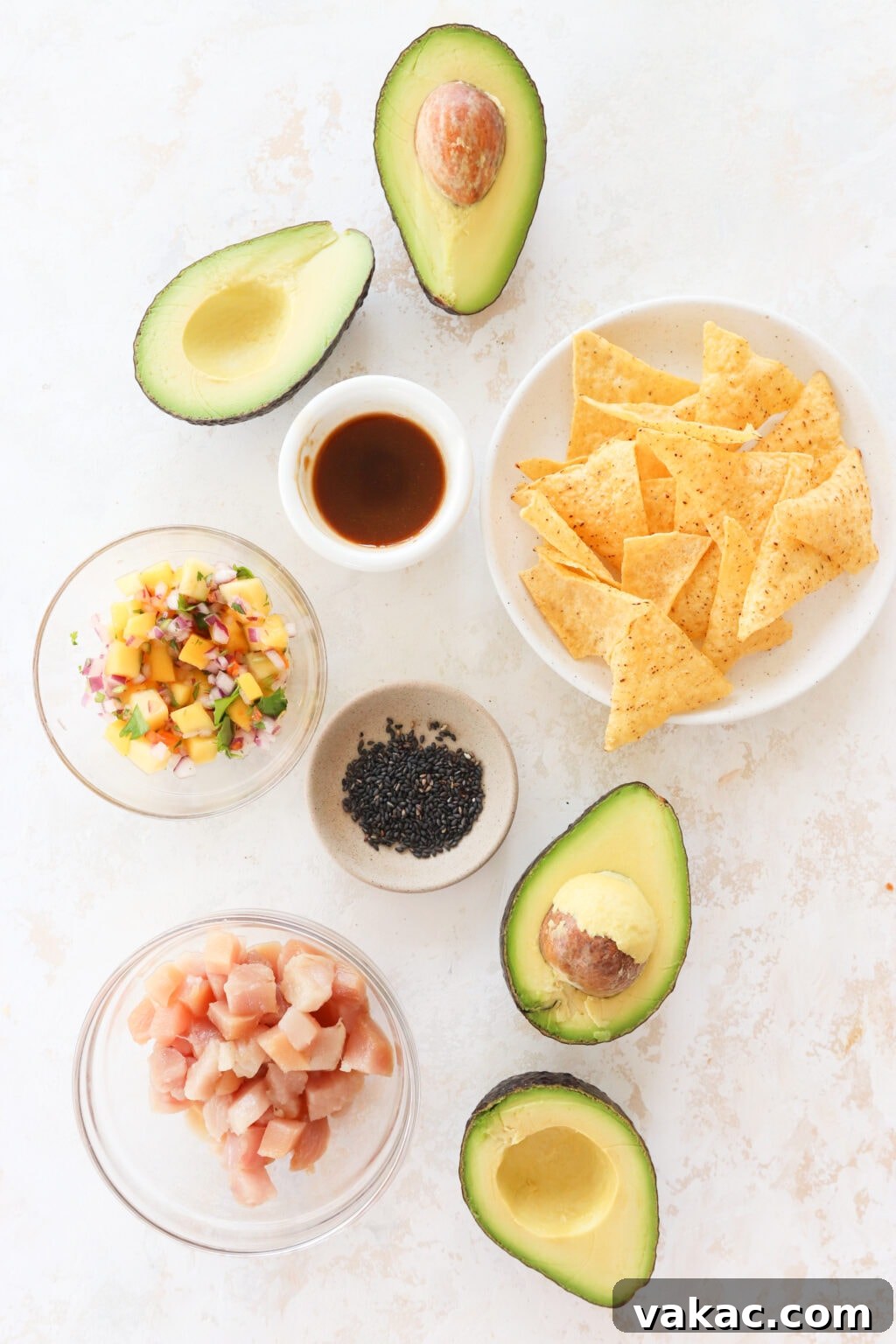 An overhead shot of ingredients for tuna avocado boats. There is an avocado cut in half, a small bowl with soy sauce, a plate of tortilla chips, a clear bowl with mango habanero salsa, a brown bowl with black sesame seeds, a clear bowl with tuna and another avocado cut into half.