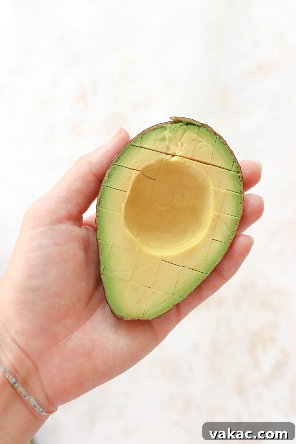 A hand is holding half an avocado. The avocado half has its skin on and has been sliced into 1/2 inch cubes.