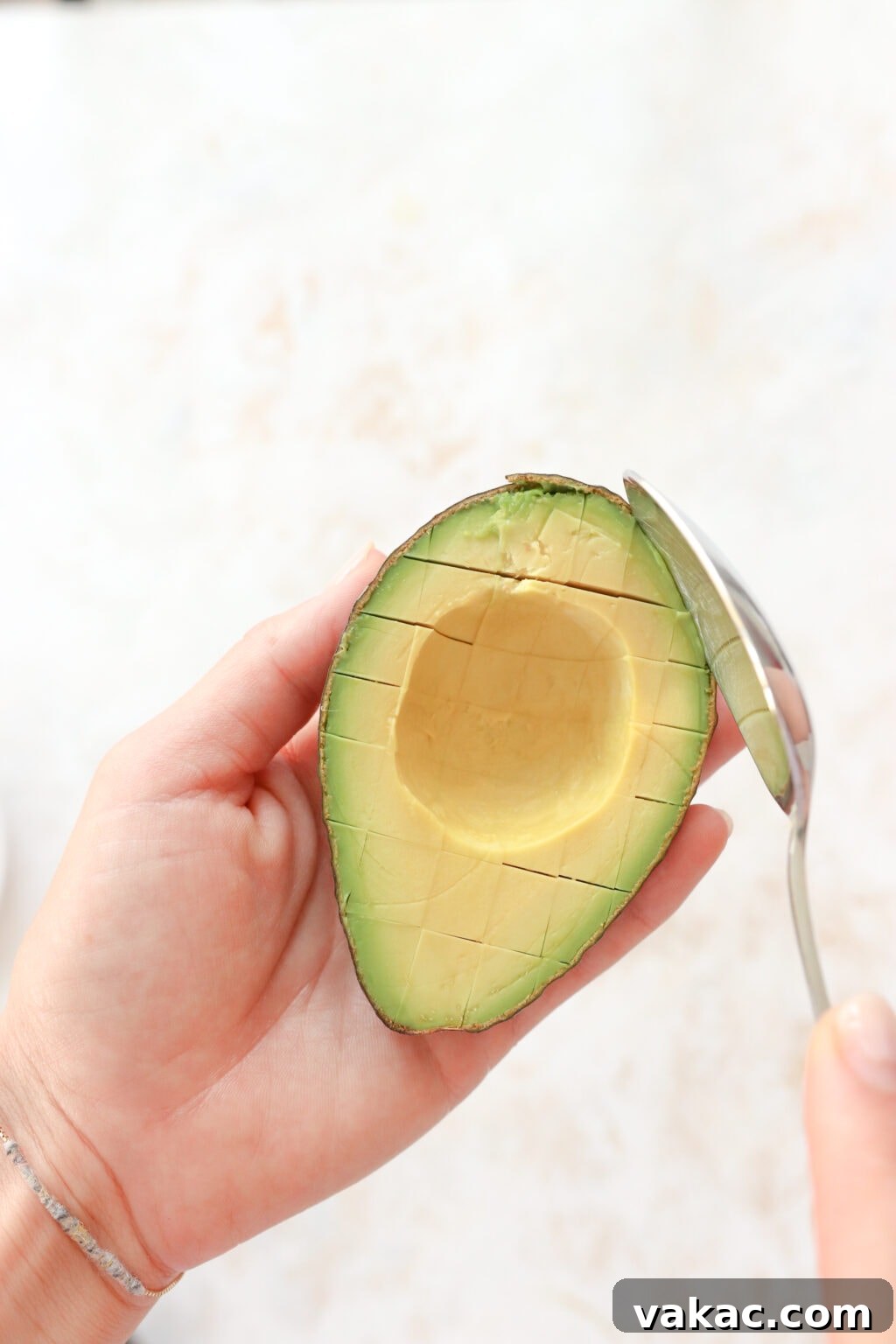 A hand is holding a scored avocado with the skin on and is using a silver spoon to scoop out the rest of the avocado.