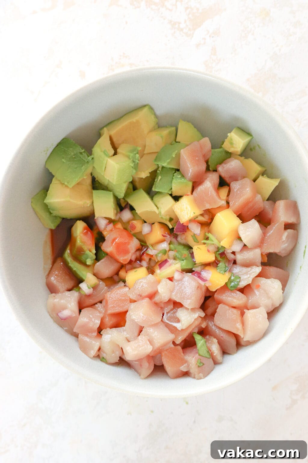 A white bowl has the ingredients for tuna avocado boats in it. At the top is cubed avocado, then mango habanero salsa, then cubed sushi-grade tuna (which is pink), and then soy sauce.