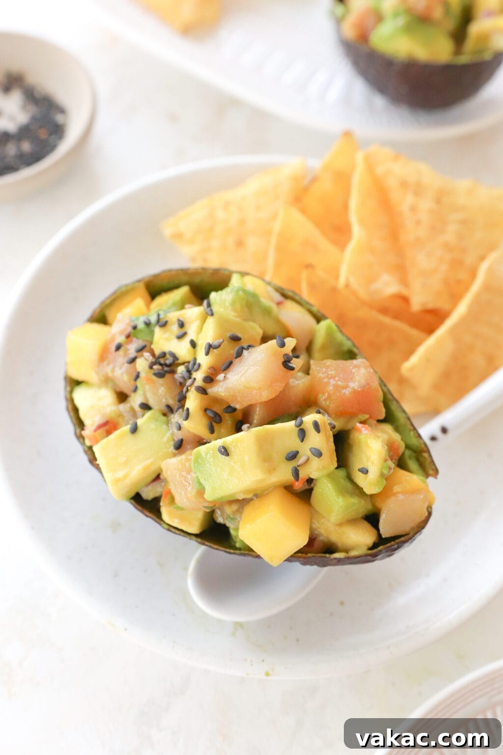 A white plate with an avocado shell filled with cubed avocado, mango and habanero salsa, and cubed tuna. Beside it are some tortilla chips and a white spoon.